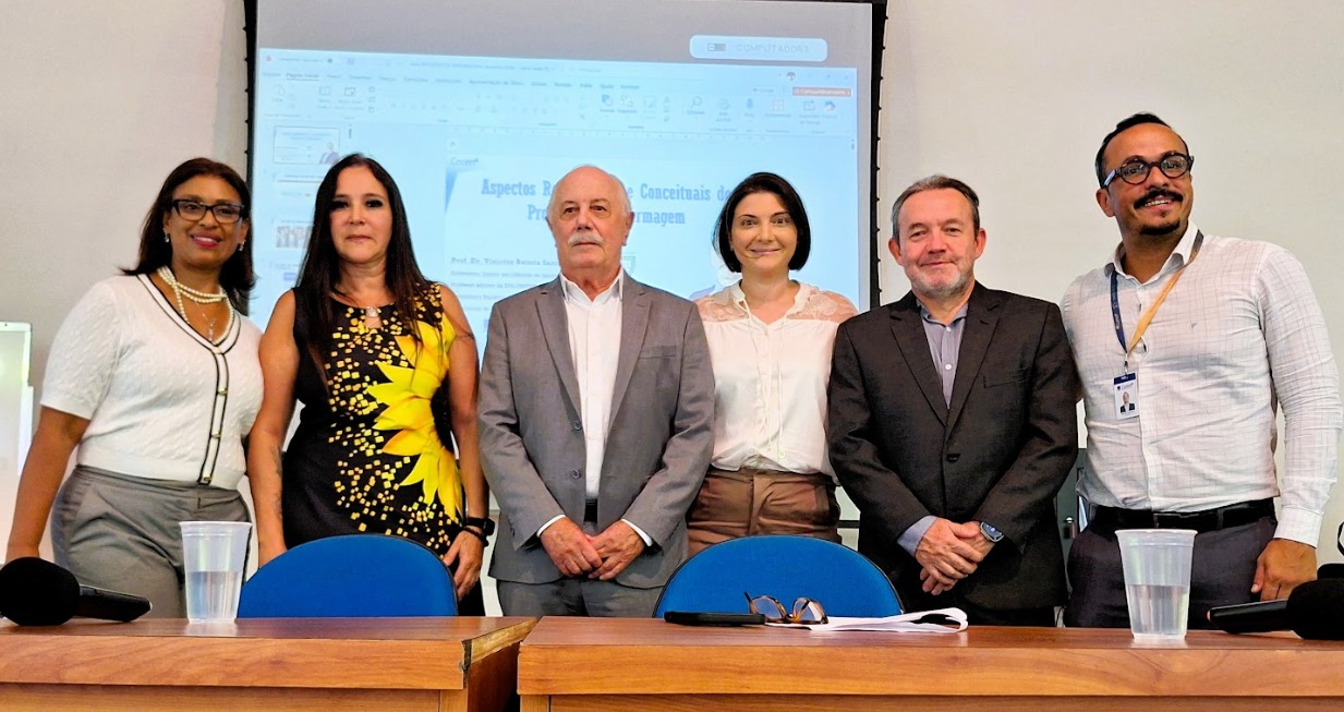 A imagem mostra seis pessoas em pé, lado a lado, posando para uma foto em um ambiente que parece ser uma sala de aula ou auditório. Elas estão atrás de duas mesas de madeira com cadeiras azuis, onde há alguns objetos como copos de plástico, um microfone e óculos. Ao fundo, há um projetor exibindo um slide de apresentação — aparentemente relacionado a um tema técnico ou acadêmico, possivelmente na área de enfermagem. O grupo é composto por quatro mulheres e dois homens, todos vestidos de forma profissional ou semiformal. No centro, um homem mais velho, de terno cinza, ocupa posição de destaque. Os demais estão distribuídos ao redor, alguns sorrindo levemente, outros com expressão neutra.