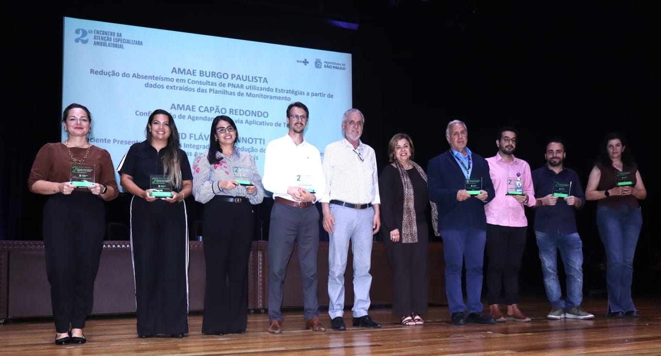 Fotografia colorida, em plano médio e horizontal, tirada em um palco de teatro durante o 2º Encontro da Atenção Especializada Ambulatorial. No centro do palco, dez pessoas (cinco mulheres e cinco homens) estão posicionadas em linha, voltadas para a câmera. Sete dessas pessoas seguram placas de premiação transparentes com detalhes em verde. Ao fundo, um grande telão exibe o logotipo do evento e o título