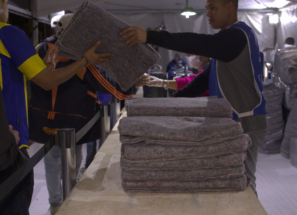 A imagem mostra uma cena de distribuição de cobertores em um ambiente que aparenta ser uma estrutura montada para atendimento emergencial ou ação social, como uma tenda. No primeiro plano, vemos uma pilha de cobertores dobrados sobre uma mesa. À direita, um homem usando colete azul entrega um cobertor a uma pessoa do outro lado da mesa. Outras pessoas com coletes e equipamentos de proteção, como máscaras, também estão envolvidas na ação