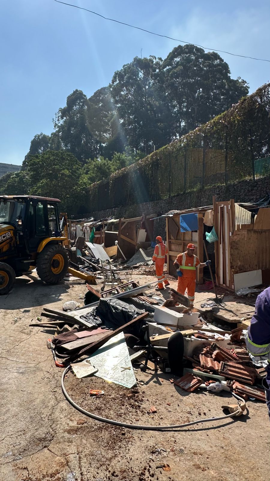 Imagem: Uma foto tirada durante o dia, mostrando uma área ao ar livre que parece ter sido um acampamento ou uma favela em processo de demolição. A luz do sol está forte, indicando um dia claro e ensolarado. Elementos principais: Primeiro plano: Uma grande quantidade de entulho e detritos espalhados pelo chão de concreto. Há pedaços de madeira, telhas, objetos metálicos retorcidos e outros materiais de construção quebrados. Um longo tubo preto de borracha está enrolado no chão. Meio do plano: Dois homens vestindo uniformes laranja com listras reflexivas estão no centro da imagem. Eles parecem estar trabalhando na limpeza ou demolição do local. Um deles está de costas para a câmera, enquanto o outro está de perfil. Fundo: Uma série de estruturas improvisadas, como barracos feitos de madeira e outros materiais, são visíveis. Essas estruturas estão parcialmente demolidas, com partes faltando ou quebradas. Ao fundo, há uma cerca alta de metal e árvores frondosas, sugerindo que o local pode estar próximo a uma área arborizada ou parque. Lateral esquerda: Uma retroescavadeira amarela e preta da marca JCB é parcialmente visível, indicando que maquinário pesado está sendo usado na operação. Lateral direita: Uma pequena parte de uma pessoa vestindo um uniforme azul é visível, sugerindo que há mais pessoas envolvidas na operação fora do quadro. Cores: As cores predominantes na imagem são o marrom e o cinza do entulho, o laranja dos uniformes dos trabalhadores e o verde das árvores ao fundo. O céu está claro e azul, indicando um dia ensolarado. Atmosfera: A imagem transmite uma sensação de desordem e destruição, mas também de trabalho e atividade, com os trabalhadores limpando a área. A presença da retroescavadeira sugere que a demolição ou limpeza está em andamento. Informações adicionais: A imagem foi tirada na vertical e tem uma boa iluminação, permitindo que os detalhes sejam claramente visíveis. A qualidade da foto é boa, com cores vibrantes e foco nítido.