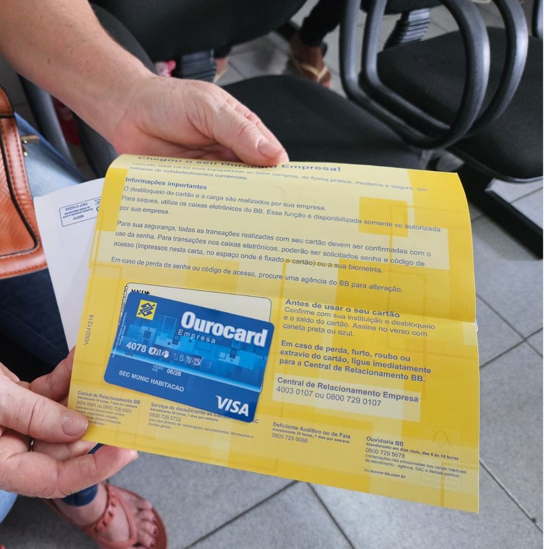 Mulher anônima mostra o cartão emergencial. Ele é azul com logotipos do Banco do Brasil e empresa de Cartões Visa. Em cima à direita está escrito Ourocard e na linha de baixo Secretaria Municipal de Habitação