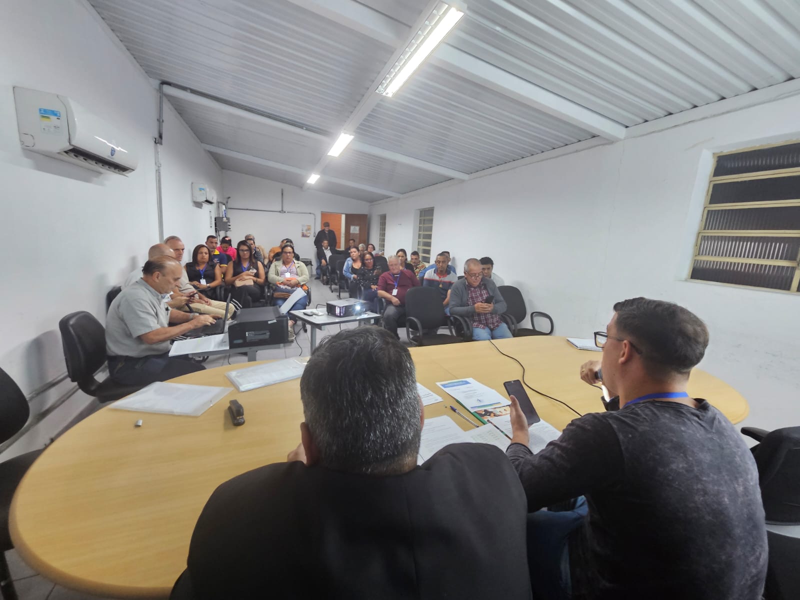 Subprefeito Eduardo Valentim ao lado do Conselheiro Rodolfo Coimbra, que está com um microfone na mão sob uma mesa amarela. No fundo, estão as pessoas presentes na reunião.