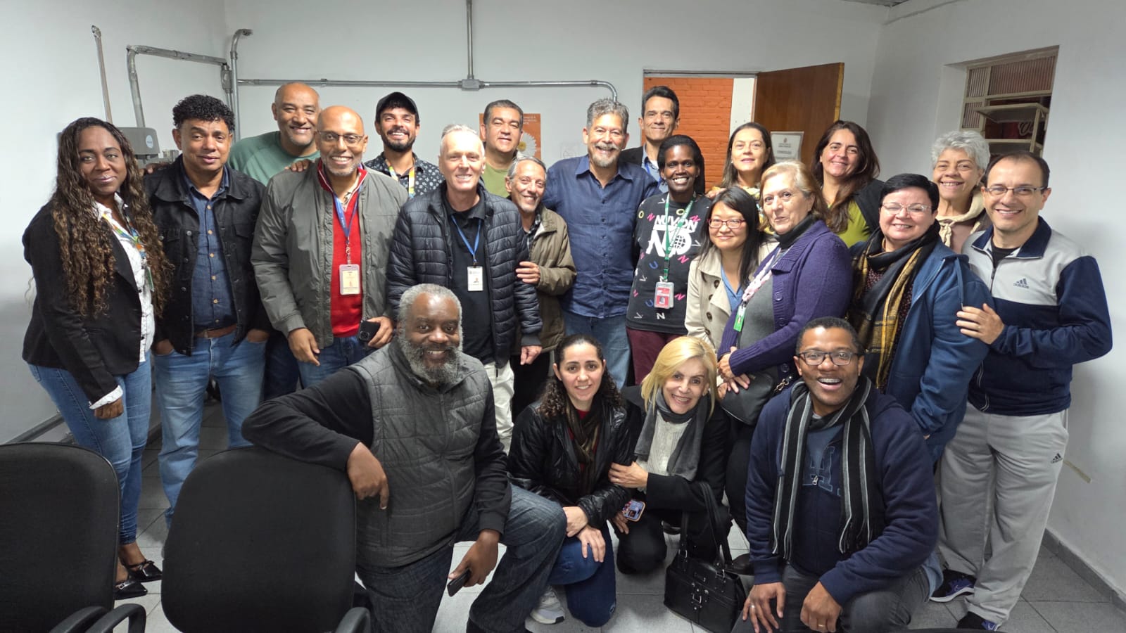 A imagem mostra um grupo diverso de aproximadamente 25 pessoas reunidas em uma sala com paredes brancas e iluminação fluorescente. Estão todas sorrindo e voltadas para a câmera, demonstrando alegria e descontração. A maioria das pessoas está de pé, e algumas estão agachadas ou ajoelhadas na frente para caberem na foto. O grupo é composto por homens e mulheres de diferentes idades, tons de pele e estilos, o que evidencia a diversidade. Algumas pessoas usam crachás no pescoço, indicando que possivelmente fazem parte de uma equipe de trabalho, projeto ou evento institucional. Ao fundo, vê-se uma porta de madeira aberta, uma janela pequena e uma parede com tubulações aparentes. O ambiente parece ser uma sala funcional, como de um escritório ou órgão público. As pessoas usam roupas casuais e de frio, como jaquetas, cachecóis e suéteres, sugerindo que a foto foi tirada em um dia de temperatura baixa. A atmosfera geral é de união, acolhimento e celebração.