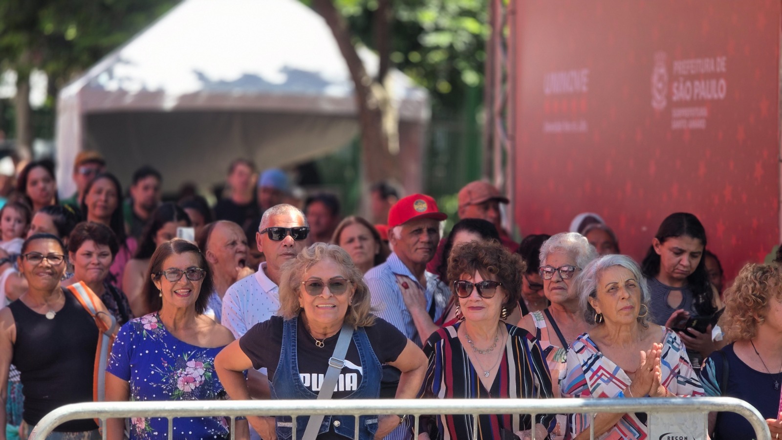 Descrição da imagem: Grupo numeroso de pessoas reunidas em um evento ao ar livre, posicionadas atrás de uma grade metálica. A maioria é composta por mulheres e idosos. Ao fundo, há uma estrutura de palco com painel vermelho contendo a identidade visual da Prefeitura de São Paulo, além de árvores e uma tenda branca.