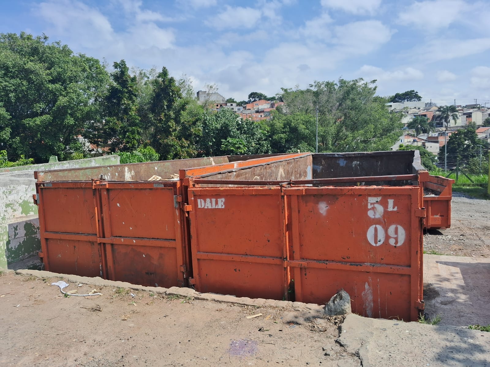 #PraCegoVer: Imagem mostra duas caçambas grandes de lixo, na cor laranja, que fazem parte de um ecoponto na zona leste de São Paulo. Ao fundo, há árvores e um terreno aberto.