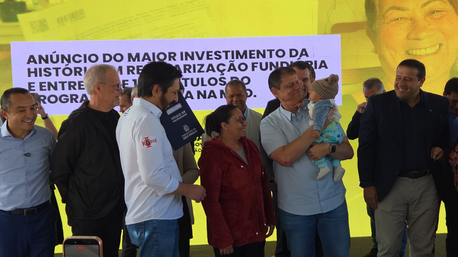A imagem mostra um grupo de pessoas reunido em um palco durante um evento público. Ao centro, um homem segura um bebê no colo e sua esposa está recebendo seu título de propriedade. Outras pessoas estão ao redor, todas de pé, participando do momento. Ao fundo, há um grande painel amarelo com texto sobre anúncio de investimentos e regularização fundiária.