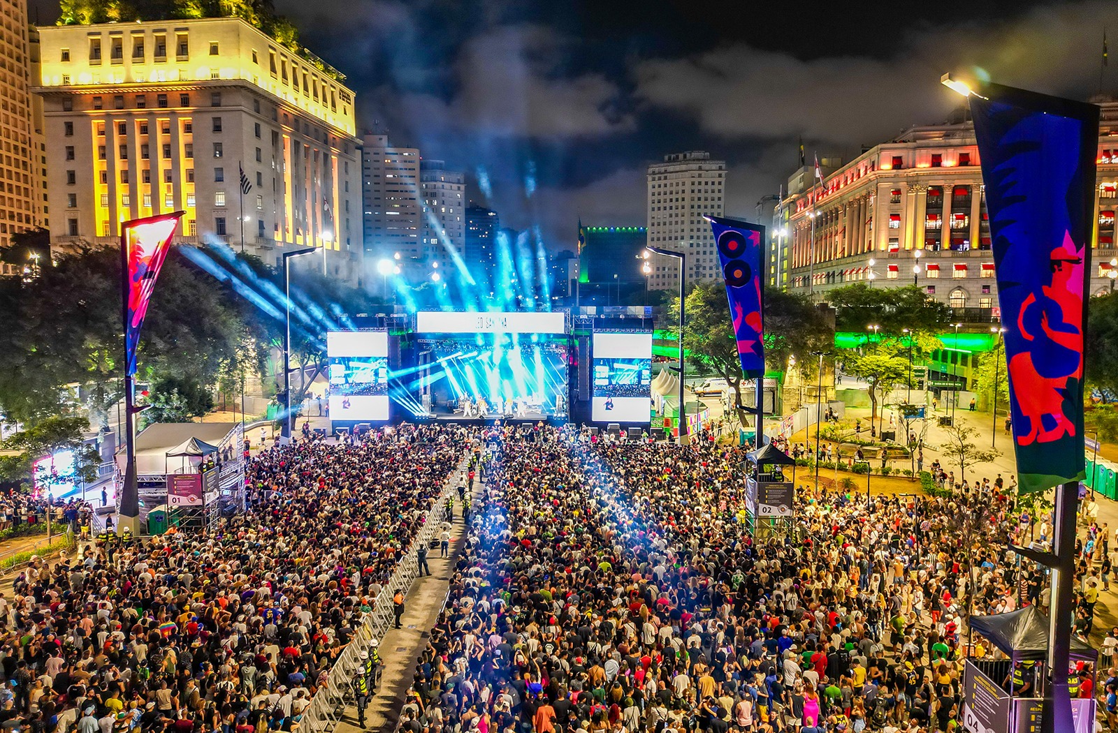 Multidão de pessoas diante de um grande palco posicionado ao fundo da imagem, durante evento no Vale do Anhangabaú, no centro da cidade de São Paulo. No canto superior direito é possível ver uma parte do prédio do Shopping Light e no canto superior esquerdo está o prédio da Prefeitura de São Paulo