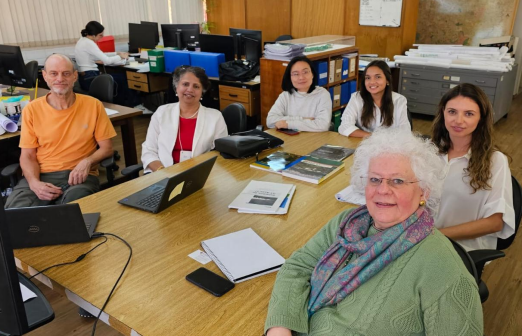 Na imagem, vemos sete pessoas reunidas ao redor de uma mesa de madeira clara em um ambiente de escritório. O grupo, composto por homens e mulheres, transmite um clima de reunião ou trabalho colaborativo. Sobre a mesa, estão laptops, cadernos, celulares e alguns documentos, sugerindo um momento de discussão ou planejamento. O ambiente tem aparência organizada, com armários, arquivos, mapas na parede e iluminação natural ao fundo. A paleta de cores é sóbria, com tons neutros predominantes, como o branco, bege, madeira e alguns toques de cor nas roupas dos presentes — como o verde e o laranja.