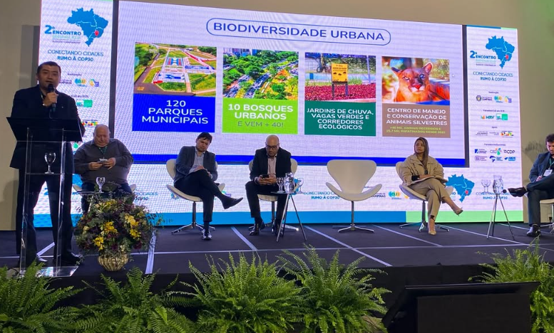 A imagem mostra um painel de debate ou apresentação sobre biodiversidade urbana, realizado em um palco com fundo digital iluminado, predominantemente em tons de azul, verde e branco. No canto esquerdo da tela, aparece o logotipo do evento com o texto "2º Encontro - Conectando Cidades rumo à COP30" acompanhado por um mapa do Brasil em azul. No palco, estão seis pessoas. Cinco delas estão sentadas em poltronas brancas de design moderno e uma pessoa está de pé, falando ao microfone em um púlpito transparente. Essa pessoa veste um paletó azul escuro, enquanto os outros participantes usam roupas formais em tons neutros, como cinza, preto e bege.  Ao fundo, uma grande tela exibe o tema da apresentação: "Biodiversidade Urbana". A projeção é colorida e dividida em blocos temáticos com textos e imagens. Entre os destaques, estão as menções a 120 parques municipais, 10 bosques urbanos com previsão de mais 40, jardins de chuva, vagas verdes e corredores ecológicos, além de um centro de manejo e conservação de animais silvestres. As cores dos blocos variam entre azul, verde claro, verde, laranja e outras tonalidades vibrantes. Algumas imagens ilustram os temas, incluindo uma vista aérea de áreas verdes, uma colmeia e um animal silvestre de pelagem alaranjada.  Na frente do palco, há arranjos de plantas naturais com folhagem verde, além de um vaso decorativo com flores amarelas e roxas. No lado direito da tela, uma coluna vertical exibe os logotipos de apoiadores e parceiros do evento, reforçando o caráter institucional e colaborativo da iniciativa.