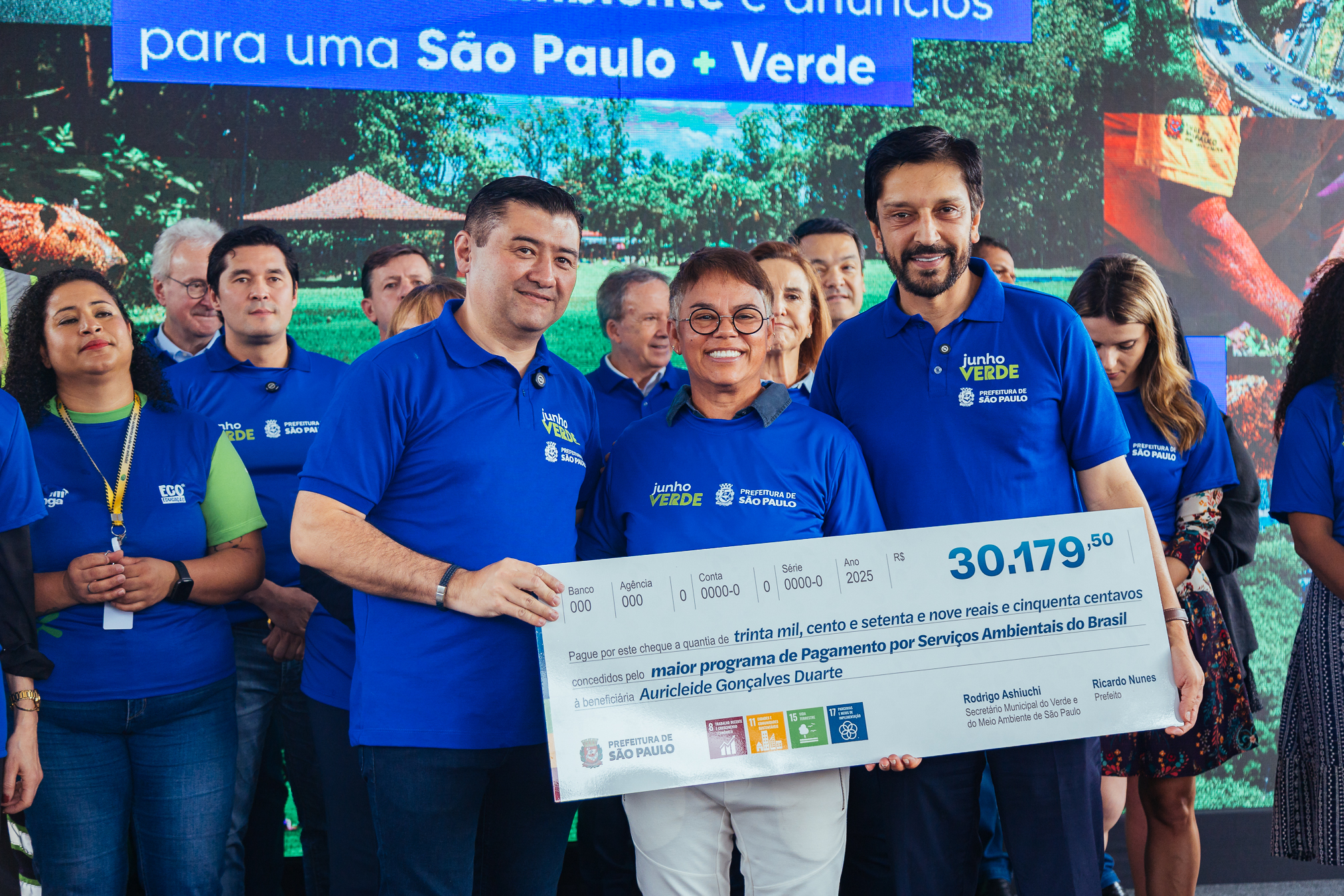 Secretário Municipal do Verde e do Meio Ambiente, Rodrigo Ashiuchi, ao lado do Prefeito Ricardo Nunes, com um dos produtores rurais beneficiado pelo Programa de Pagamento por Serviços Ambientais (PSA Mananciais). Eles posam para foto juntos atrás de um banner do evento do Dia do Meio Ambiente, que aconteceu no Parque Independência. 
