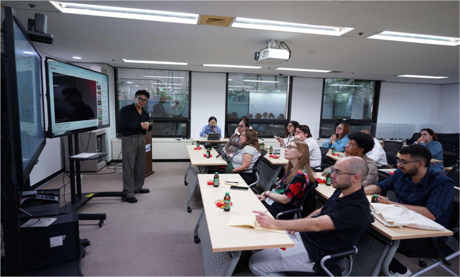 Pessoas sentadas em cadeiras escolares, dentro de uma sala de aula, assistem a uma apresentação exibida em uma televisão. Um homem está de pé, explicando o conteúdo mostrado na tela.
