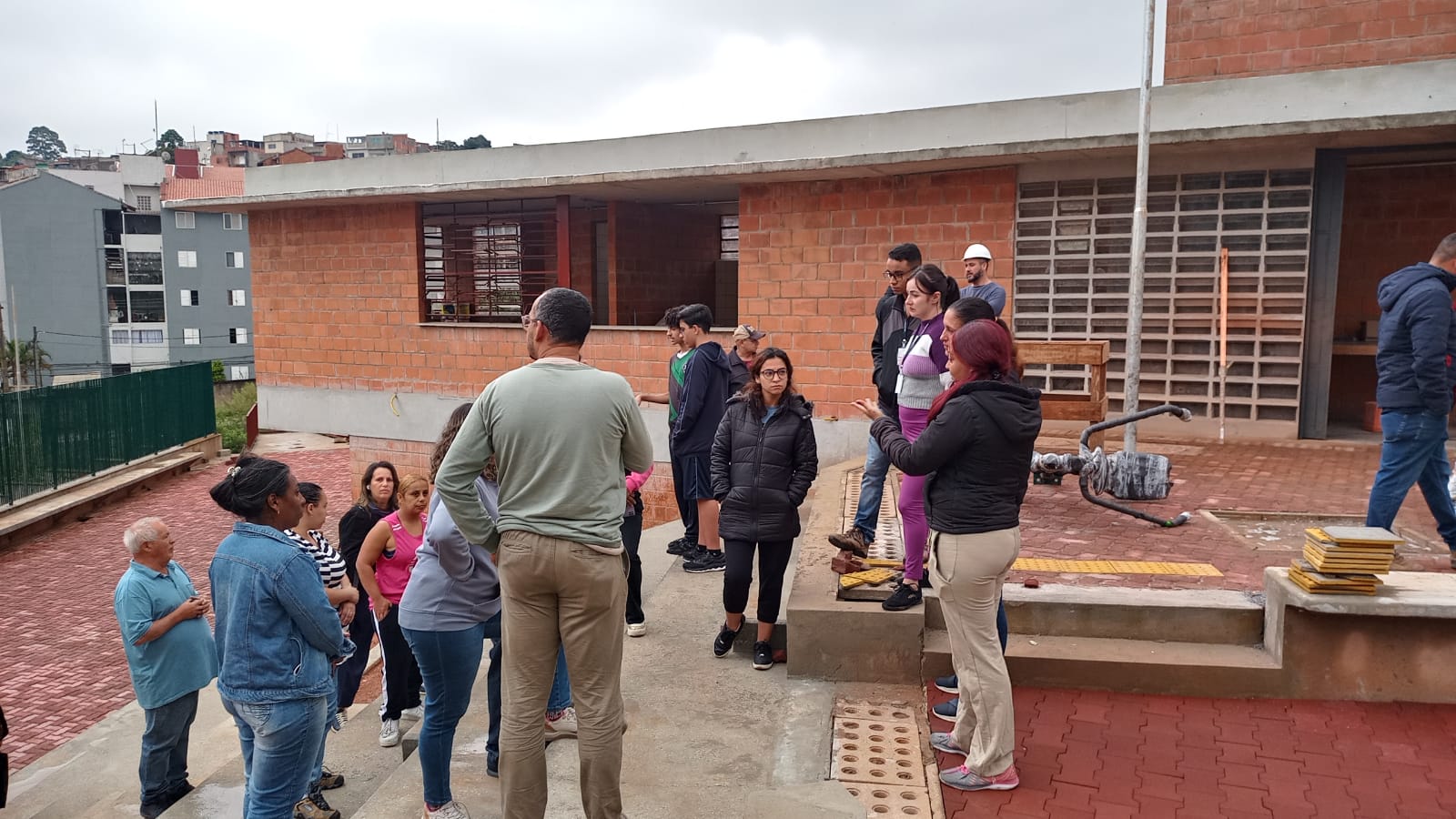 A imagem mostra um grupo de pessoas reunidas em um espaço externo de um prédio em construção. As paredes do prédio ao fundo são de tijolos aparentes em tom alaranjado, sem acabamento. Há uma calçada de piso intertravado vermelho e algumas partes de concreto cinza.  O céu está nublado, com tonalidade cinza clara. As pessoas estão usando roupas variadas, com cores como azul, cinza, preto, verde, lilás, roxo e bege. Algumas estão de jaquetas, indicando que o clima está frio. No canto direito da imagem, há uma pilha de pisos táteis amarelos. Um equipamento metálico com pintura desgastada aparece no chão próximo a um poste.  Ao fundo, do lado esquerdo, é possível ver prédios residenciais de diferentes cores: cinza, verde, branco e vermelho, compondo o cenário urbano do entorno.