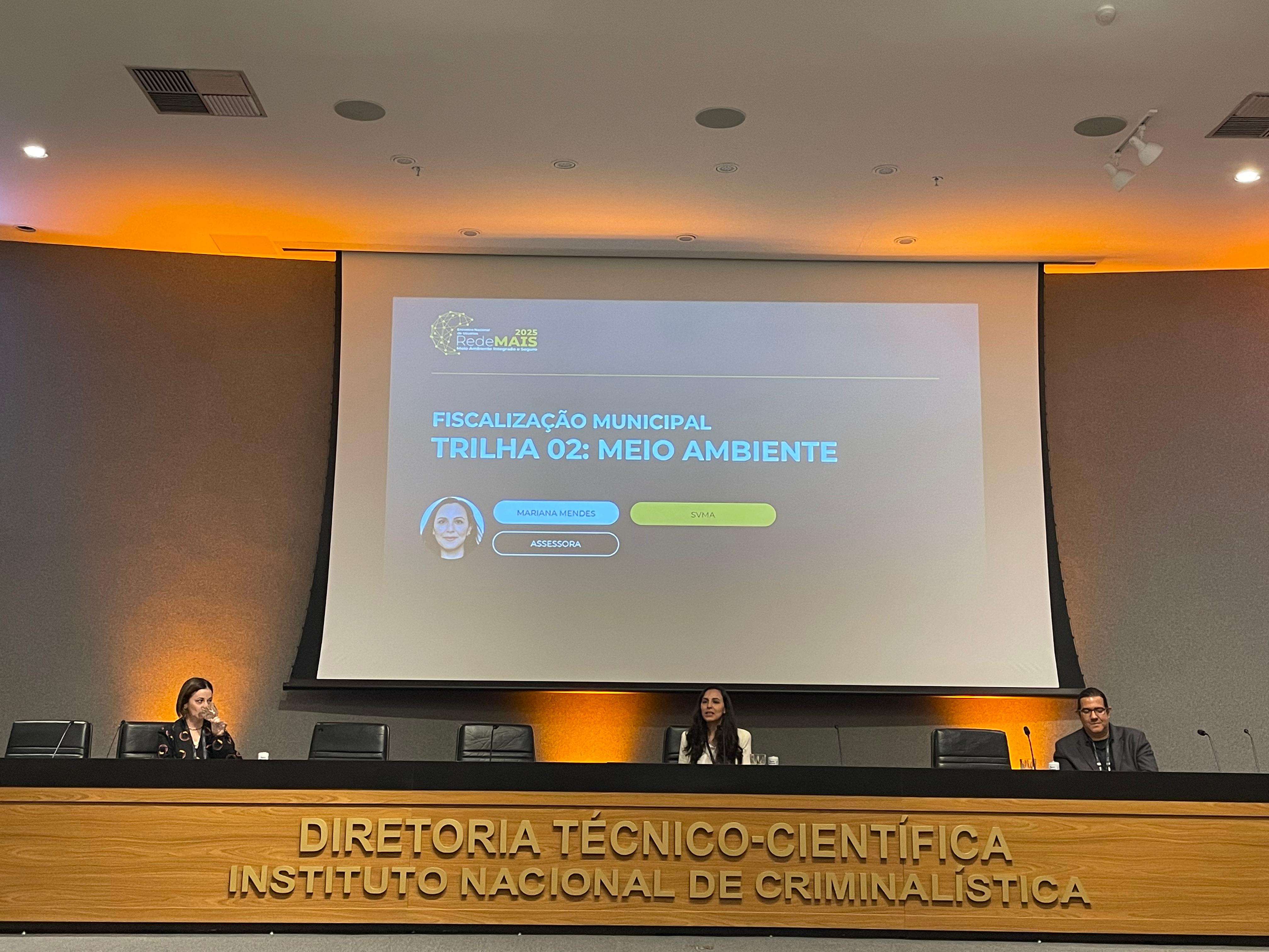 A imagem mostra um auditório durante uma apresentação. Ao fundo, há uma grande tela de projeção com fundo cinza escuro e detalhes em amarelo e azul. O título projetado diz: "Fiscalização Municipal - Trilha 02: Meio Ambiente", com o nome da palestrante, Mariana Mendes, identificada como assessora da SVMA (Secretaria do Verde e do Meio Ambiente). No canto superior esquerdo da projeção, aparece o logotipo do evento: Rede MAIS 2025.  Três pessoas estão sentadas à mesa de frente para a plateia: uma mulher à esquerda, outra mulher ao centro e um homem à direita. O fundo da parede é cinza escuro, com iluminação âmbar (alaranjada) nas laterais inferiores, criando um efeito de luz quente no ambiente.  Na frente da mesa dos palestrantes, há uma faixa de madeira clara com a inscrição em letras metálicas: "Diretoria Técnico-Científica - Instituto Nacional de Criminalística".  As cores predominantes da imagem são: cinza, preto, madeira clara (amarelo claro) e laranja suave da iluminação.