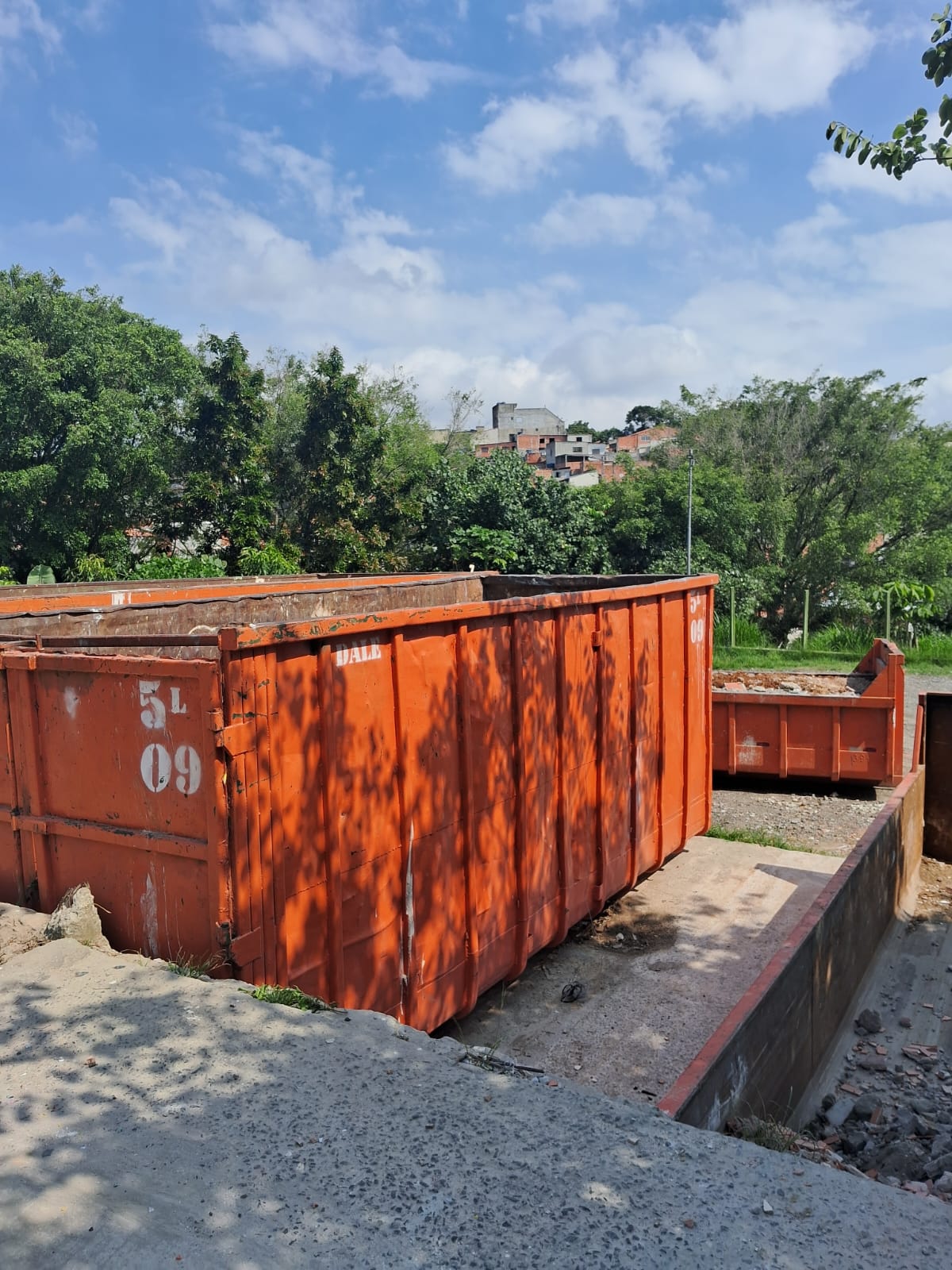 #PraCegoVer: Imagem mostra duas caçambas grandes de lixo, na cor laranja, que fazem parte de um ecoponto na zona leste de São Paulo. Ao fundo, há árvores e um terreno aberto.