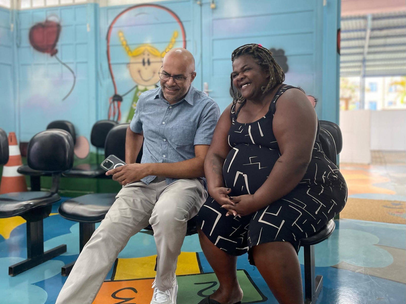 #PraTodosVerem: Subprefeito Ozziel Souza conversa e dá risadas ao lado de uma munícipe. Ambos estão sentados. Ozziel veste uma camisa social na cor azul e a mulher está usando um vestido preto.
