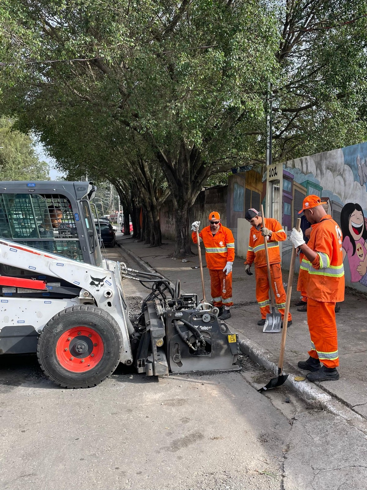 #PraTodosVerem: Três homens da equipe de zeladoria da Subprefeitura São Mateus estão fazendo uma ação de tapa-buraco na rua. Eles usam uniformes na cor laranja. Junto a eles, está um caminhão tapa-buraco auxiliando o serviço.