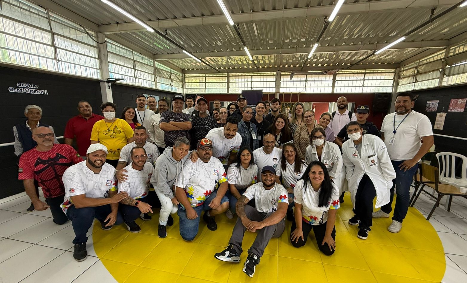 Espaço iluminado, paredes pretas com janelas, o chão branco com um grande círculo amarelo pintado. Diversas pessoas reunidas utilizando camiseta branca e sorrindo para a foto. No canto esquerdo, duas pessoas vestindo camiseta vermelha e uma pessoa de amarelo.