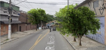 Foto de uma rua em São Paulo, Brasil, chamada Rua Nova Brasil. A rua é asfaltada e tem uma faixa amarela no centro. Há construções de um e dois andares em ambos os lados da rua, algumas com fachadas simples e outras com grafites. Uma mulher com uma mochila rosa está caminhando na calçada à direita. Um carro preto e uma motocicleta estão dirigindo na rua, em direção à câmera. Fios elétricos cruzam o céu nublado. Há árvores e vegetação visíveis entre as construções. A foto parece ter sido tirada durante o dia, sob luz natural difusa.