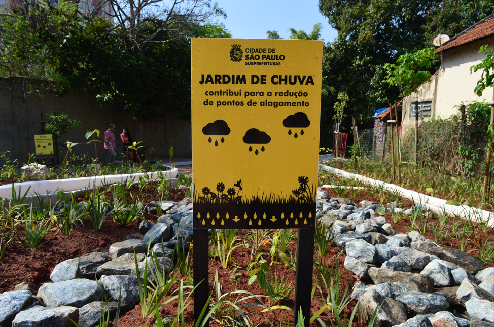 Foto de um jardim de chuva, uma área ajardinada projetada para absorver a água da chuva. O jardim possui depressões no terreno cobertas por vegetação rasteira e densa, com algumas plantas mais altas e árvores ao fundo. Há estruturas de concreto em formato de "U" invertido, com aberturas que parecem direcionar o fluxo da água. Uma área de terra batida e grama rala precede o jardim de chuva. Ao fundo, observa-se mais vegetação e a silhueta de prédios na paisagem urbana. Uma placa com o texto "CIDADE DE SÃO PAULO SUBPREFEITURAS JARDIM DE CHUVA contribui para a redução de pontos de alagamento" é parcialmente visível no canto superior esquerdo, indicando a localização e a função do jardim. O céu está parcialmente nublado, com áreas de céu azul visíveis.