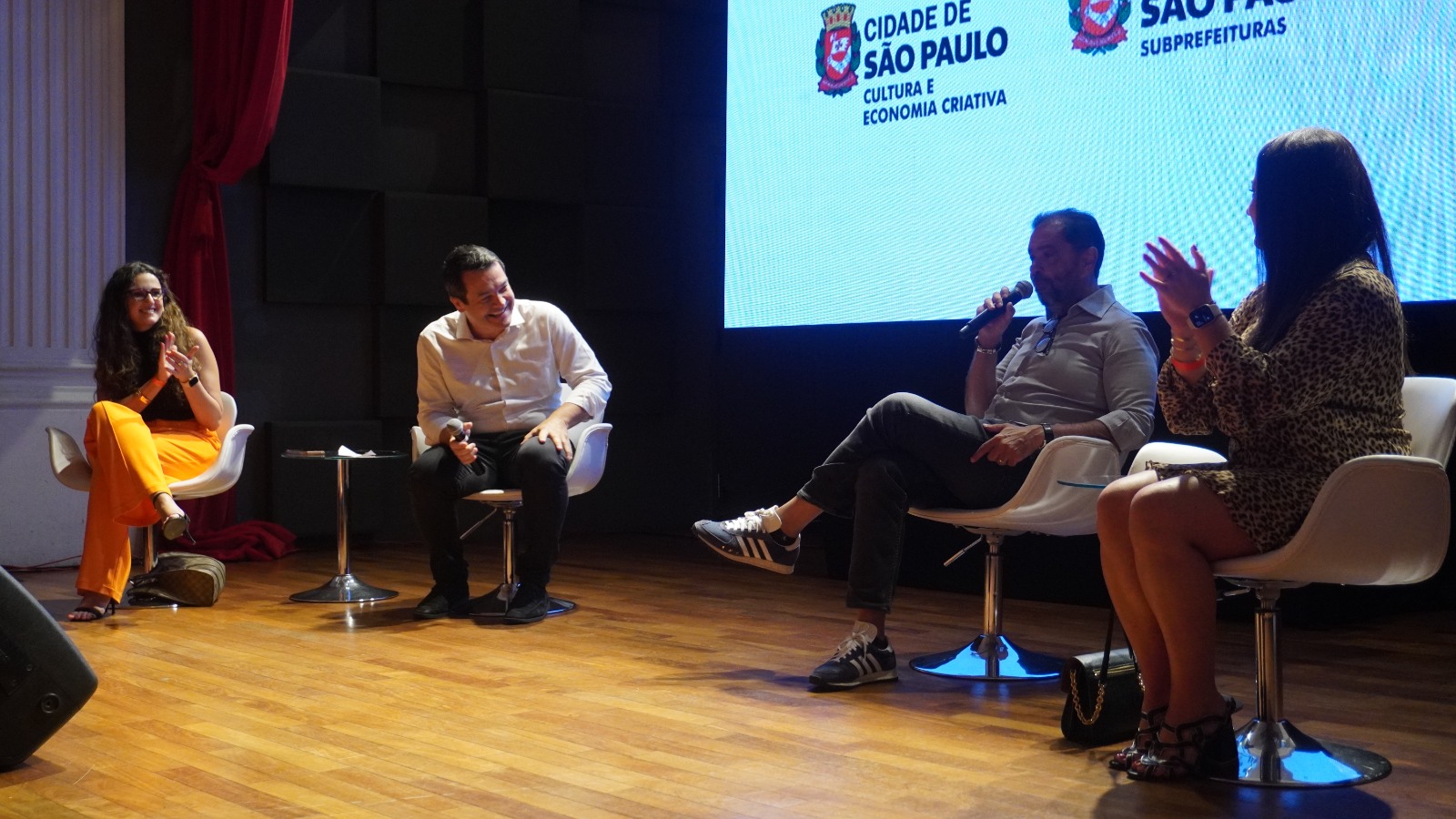 Foto de um palco com quatro pessoas sentadas em cadeiras brancas, participando de um evento. Ao fundo, uma tela de LED com o logo da "CIDADE DE SÃO PAULO". As pessoas estão vestindo roupas coloridas e modernas, e a iluminação é suave.